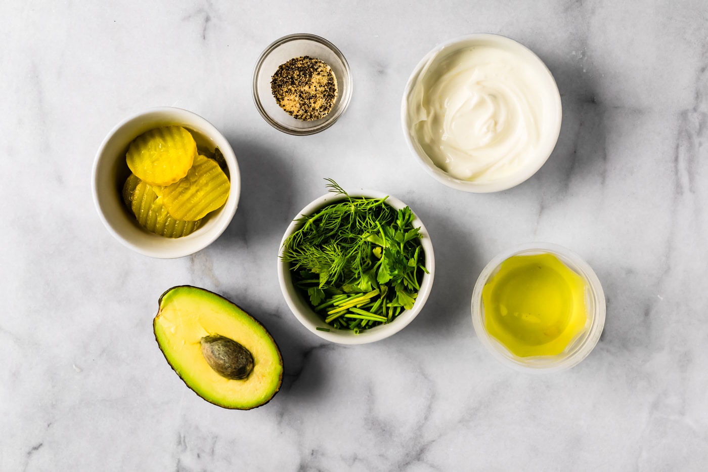 Simple ingredient setup with avocado, sliced pickles, fresh herbs, yogurt, and oil on a marble surface.