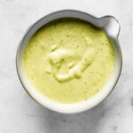 Close-up of creamy pickle dressing in a bowl, with a pale green color and specks of fresh herbs throughout.