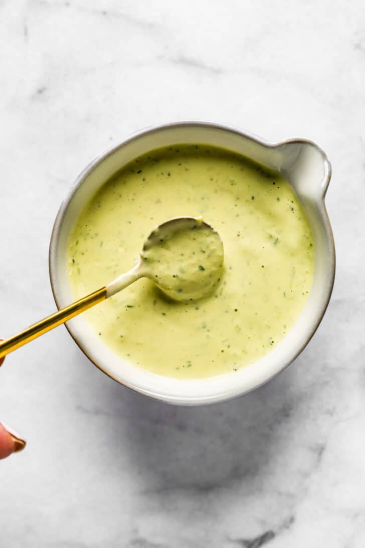 Spoon lifting thick, creamy dill pickle dressing from a small white pitcher, showing its smooth texture and green herb flecks.