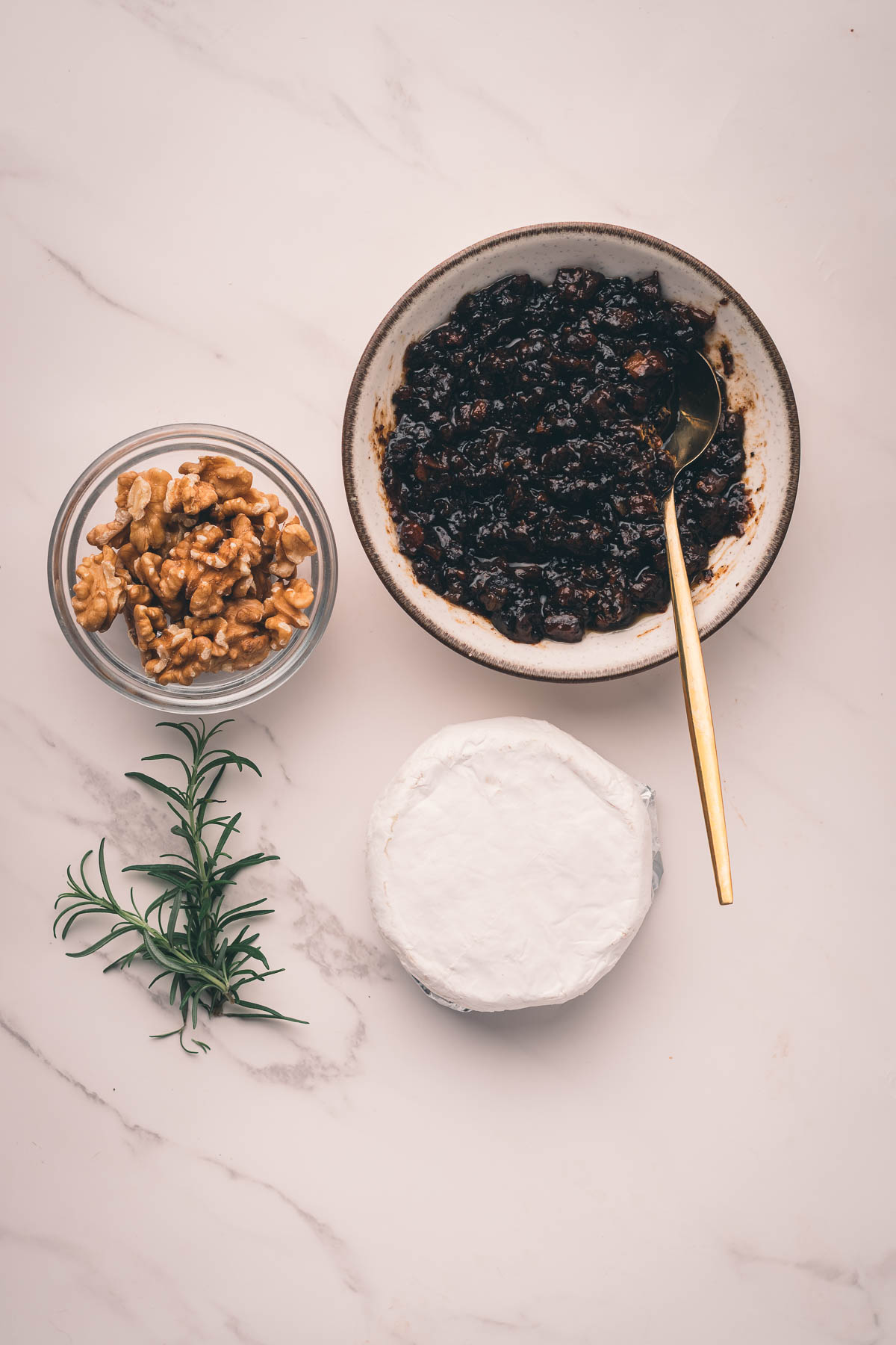 A round of Camembert cheese, a small bowl of walnuts, a sprig of rosemary, and a bowl of dark bacon jam with a gold spoon on a light marble surface.