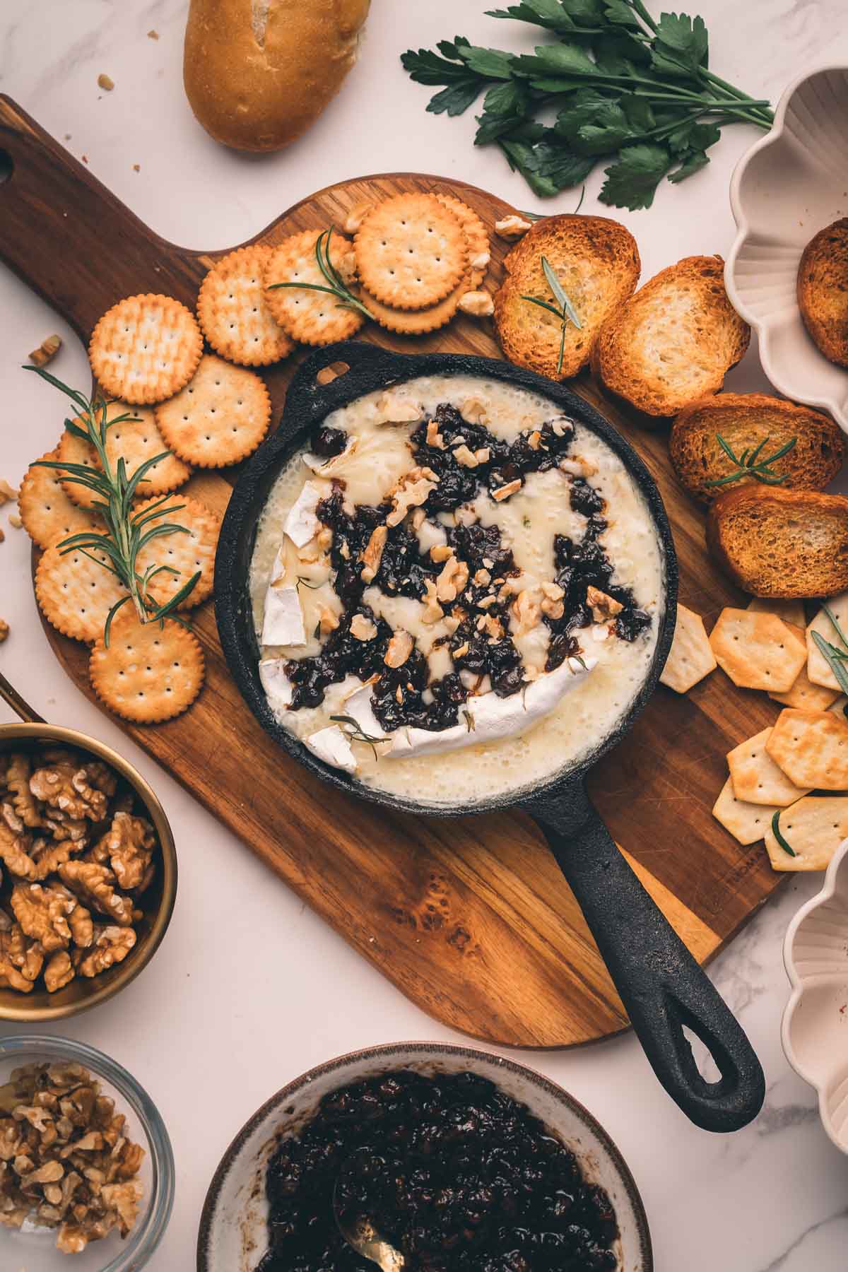 A small cast iron skillet with baked Camembert topped with walnuts and bacon jam, surrounded by crackers, toasted bread, walnuts, and parsley on a wooden board.