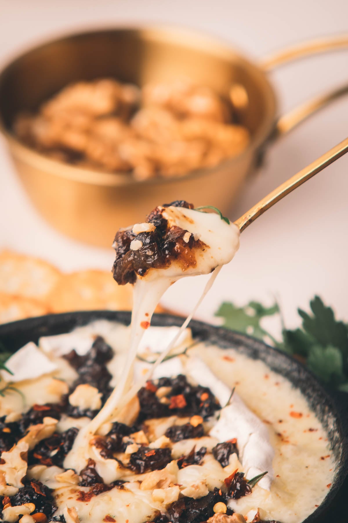 A close-up of melted cheese being lifted from a bowl topped with bacon jam, nuts, and herbs, with crackers and a bowl of food in the background.