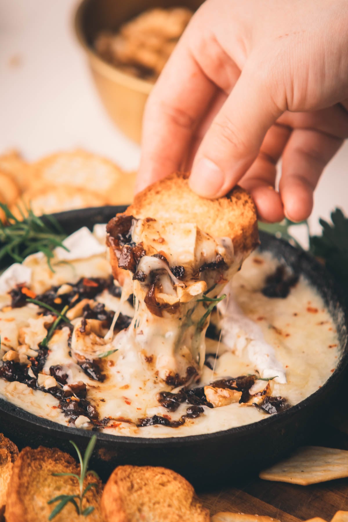 A hand dips a toasted bread slice into melted Camembert cheese topped with herbs and bacon jam.
