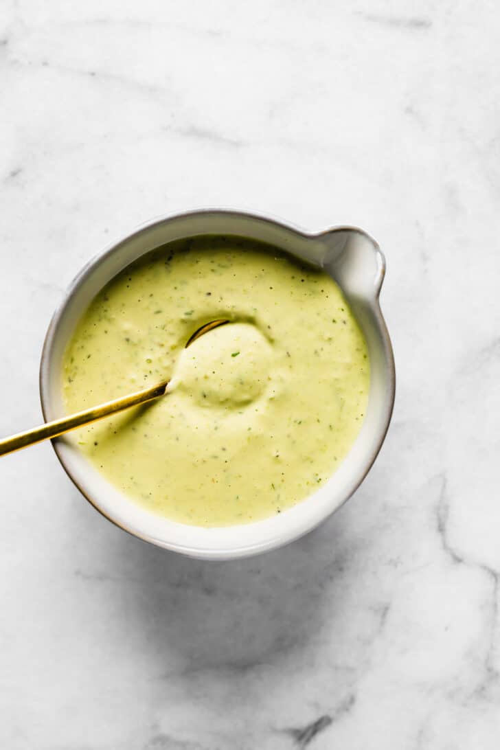 Small gold spoon sitting in a small bowl of thick creamy dill pickle dressing.