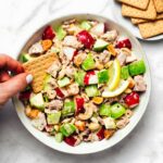 Hand holding a gluten-free cracker over a bowl of creamy tuna salad with green apple, celery, radish, cashews, and lemon wedges.