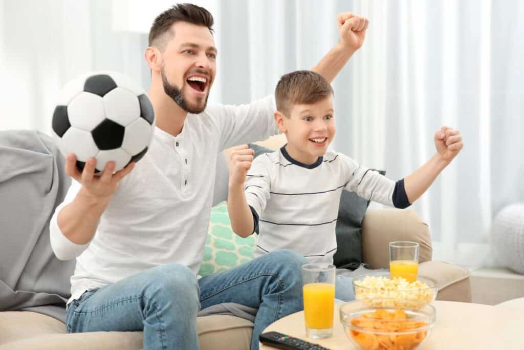 Father and son cheering while watching football at home with snacks.