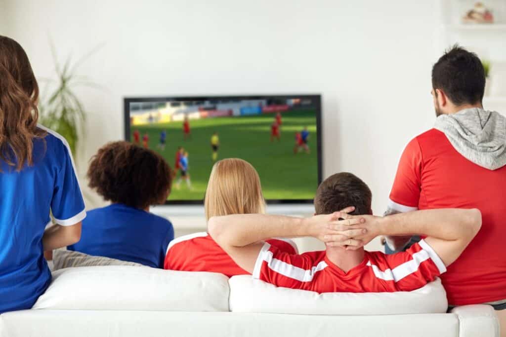 Friends sitting on a sofa watching football on TV at home.