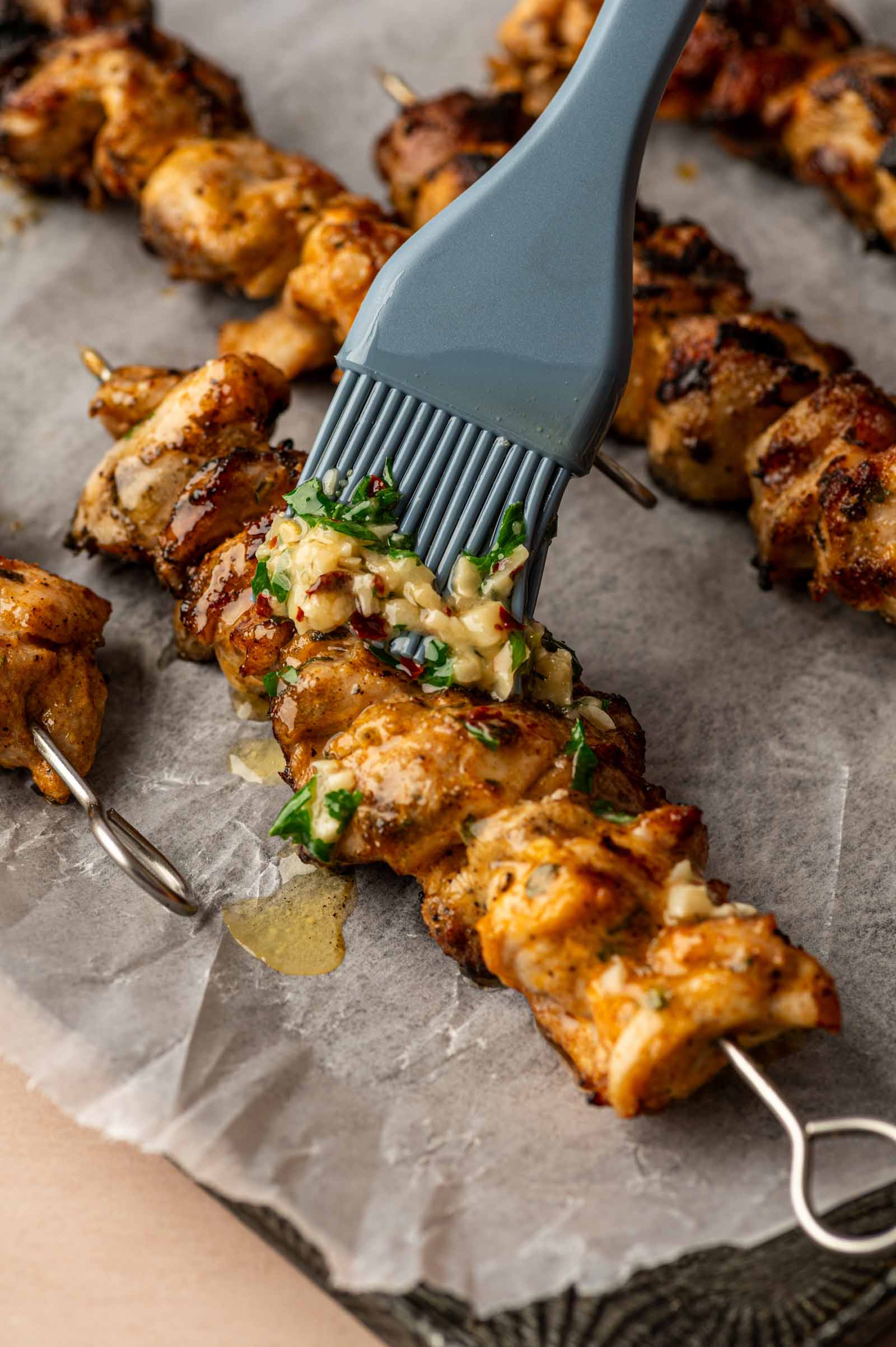A silicone brush applies a garlic parmesan sauce to grilled chicken skewers resting on parchment paper.
