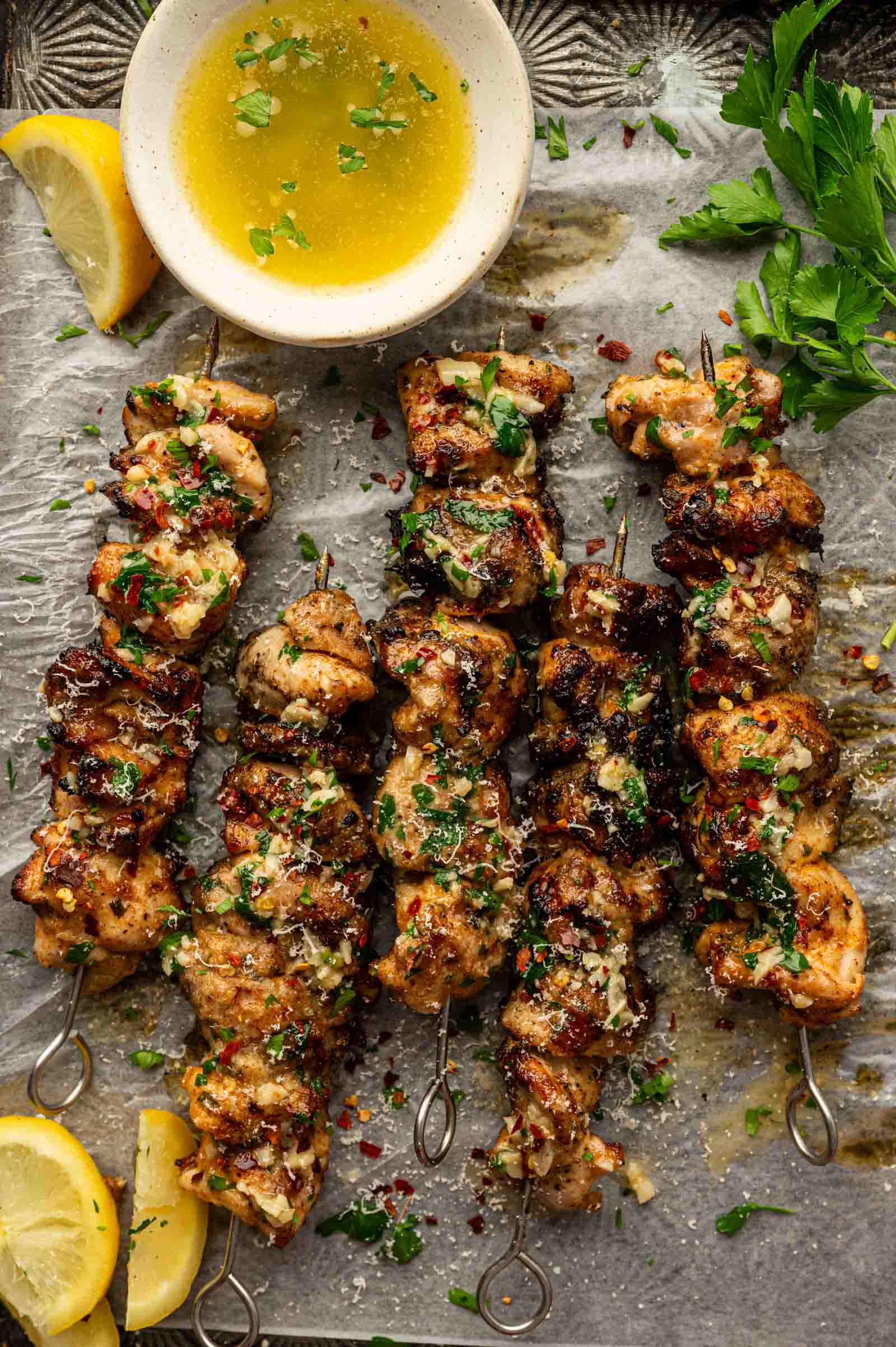 Five grilled chicken skewers drenched in garlic parmesan sauce and garnished with herbs, served with lemon wedges and a bowl of sauce on parchment paper.