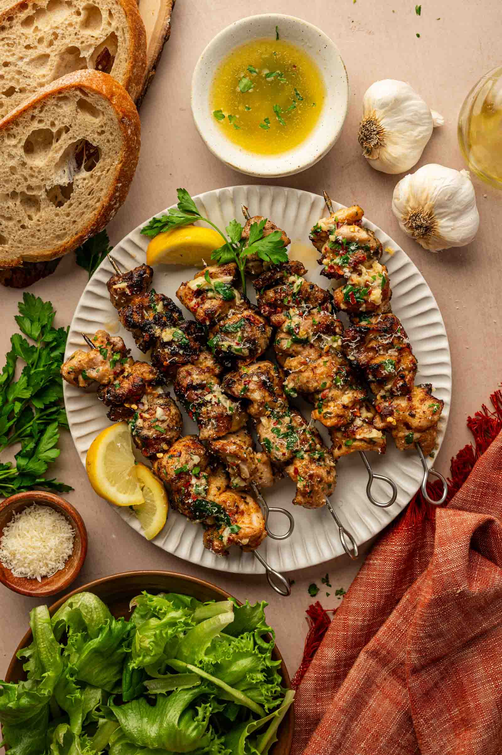 A plate of grilled garlic parmesan chicken skewers garnished with parsley and lemon slices, surrounded by bread, salad greens, garlic, grated cheese, and a bowl of garlic parmesan butter sauce.