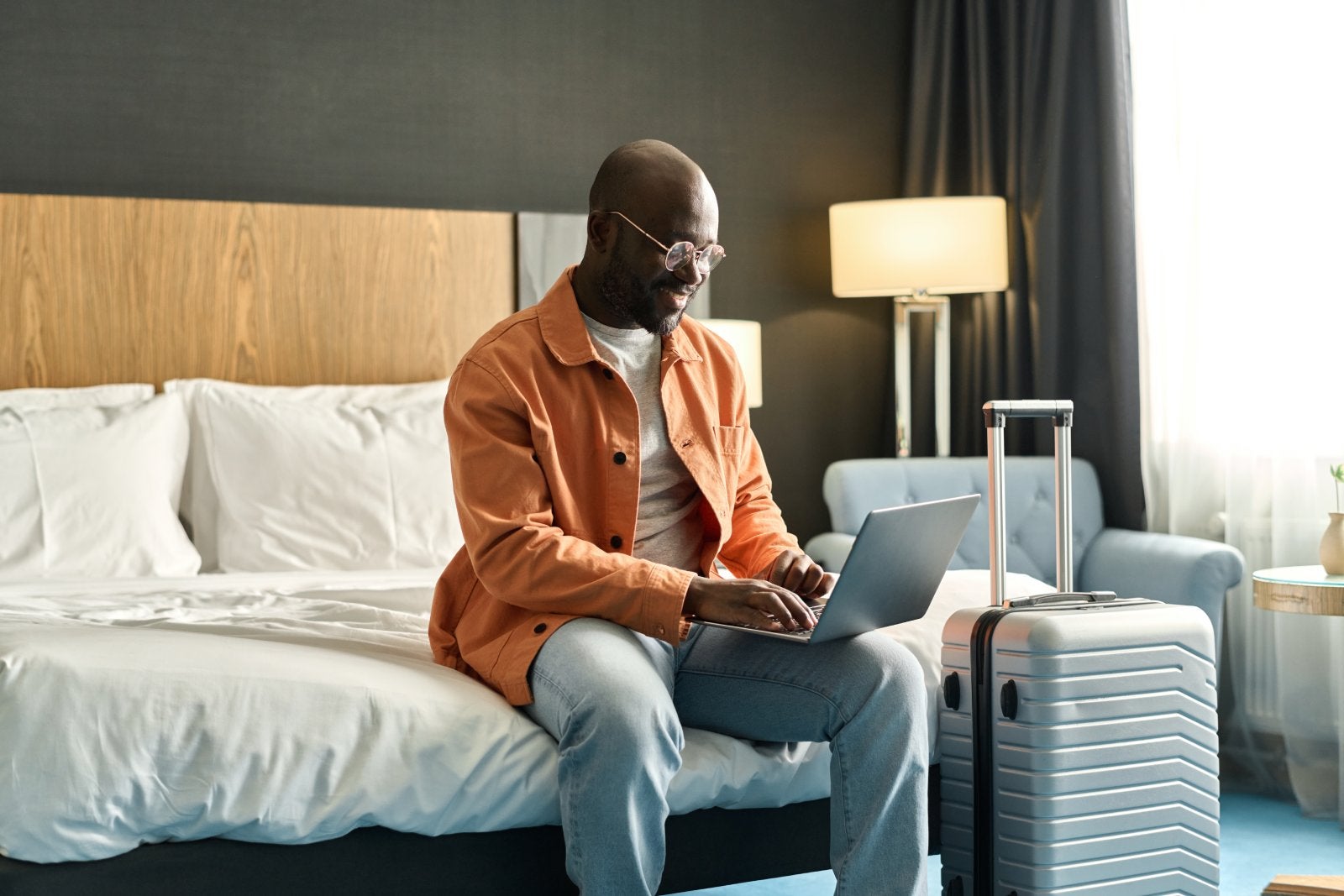 Portrait of Black man using laptop sitting on bed in hotel room and researching guest services copy space