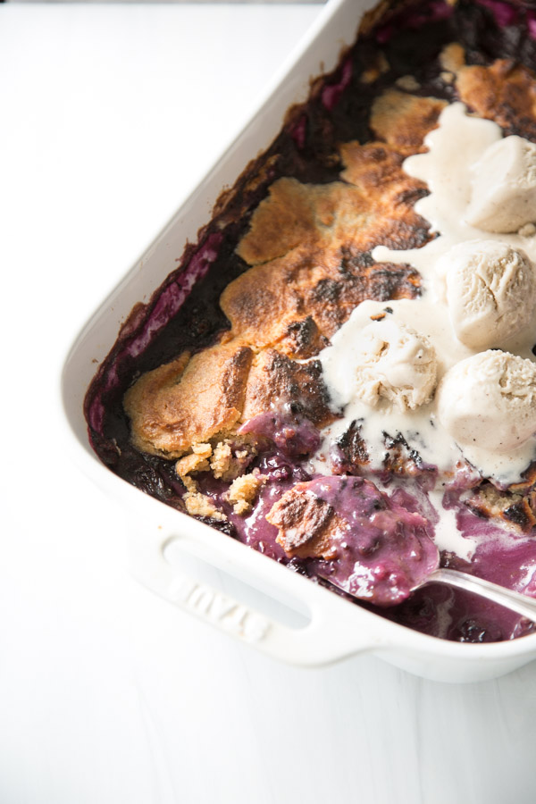 A close-up of a gluten-free blueberry cobbler in a white dish, topped with melting scoops of vanilla ice cream. The almond flour crust is golden-brown and crisp, while the bubbling berry filling is rich and purple.