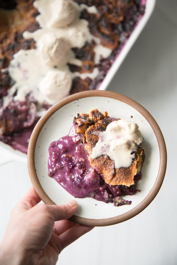 A hand holds a plate with a serving of gluten-free blackberry cobbler topped with melting vanilla ice cream; the cobbler, made with almond flour, and more scoops of ice cream are visible in the background.