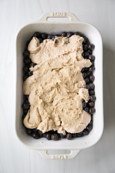 A white baking dish filled with blueberries, partially covered with dollops of almond flour biscuit dough, this gluten-free blueberry cobbler is ready to be spread and baked.