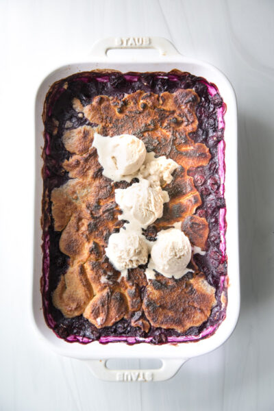 A baked blueberry cobbler with a golden-brown crust is topped with four scoops of vanilla ice cream, served in a white rectangular dish on a light marble surface—made gluten-free with almond flour.