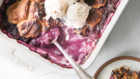 A rectangular baking dish of gluten-free blueberry cobbler with a golden-brown crust, made with almond flour, is topped with vanilla ice cream. A spoon rests inside, and a serving is plated nearby.