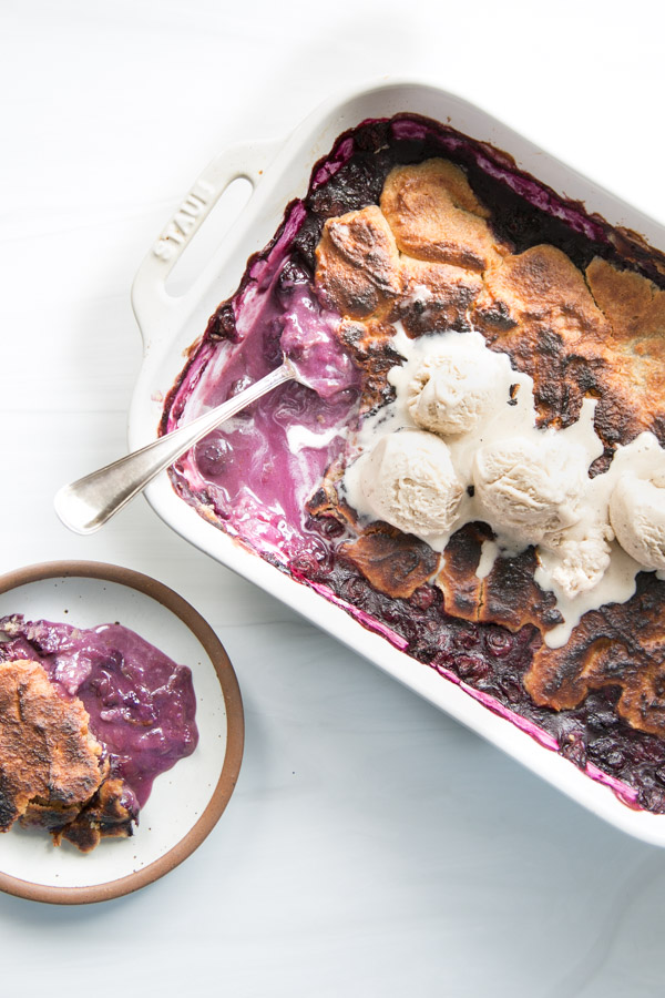 A baking dish of gluten-free blueberry cobbler made with almond flour, topped with scoops of vanilla ice cream. A serving spoon and a portion on a plate reveal the golden-brown crust and luscious purple blueberry filling.