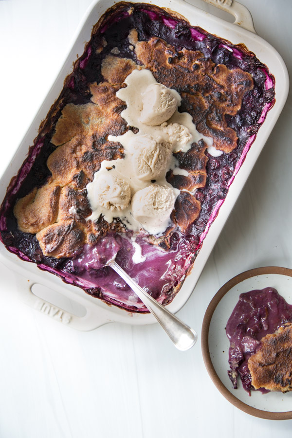A gluten-free blueberry cobbler baked in a white dish with almond flour, topped with scoops of vanilla ice cream, and served with a spoon and a portion on a small plate beside it.