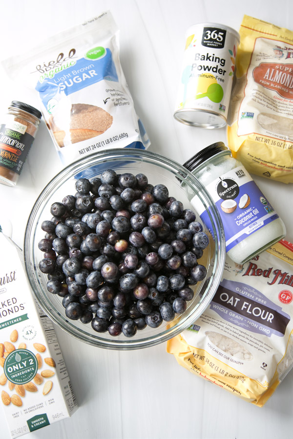 A glass bowl filled with fresh blueberries sits on a white surface, surrounded by gluten-free baking ingredients like almond flour, oat flour, coconut oil, brown sugar, baking powder, and cinnamon—perfect for a blueberry cobbler.
