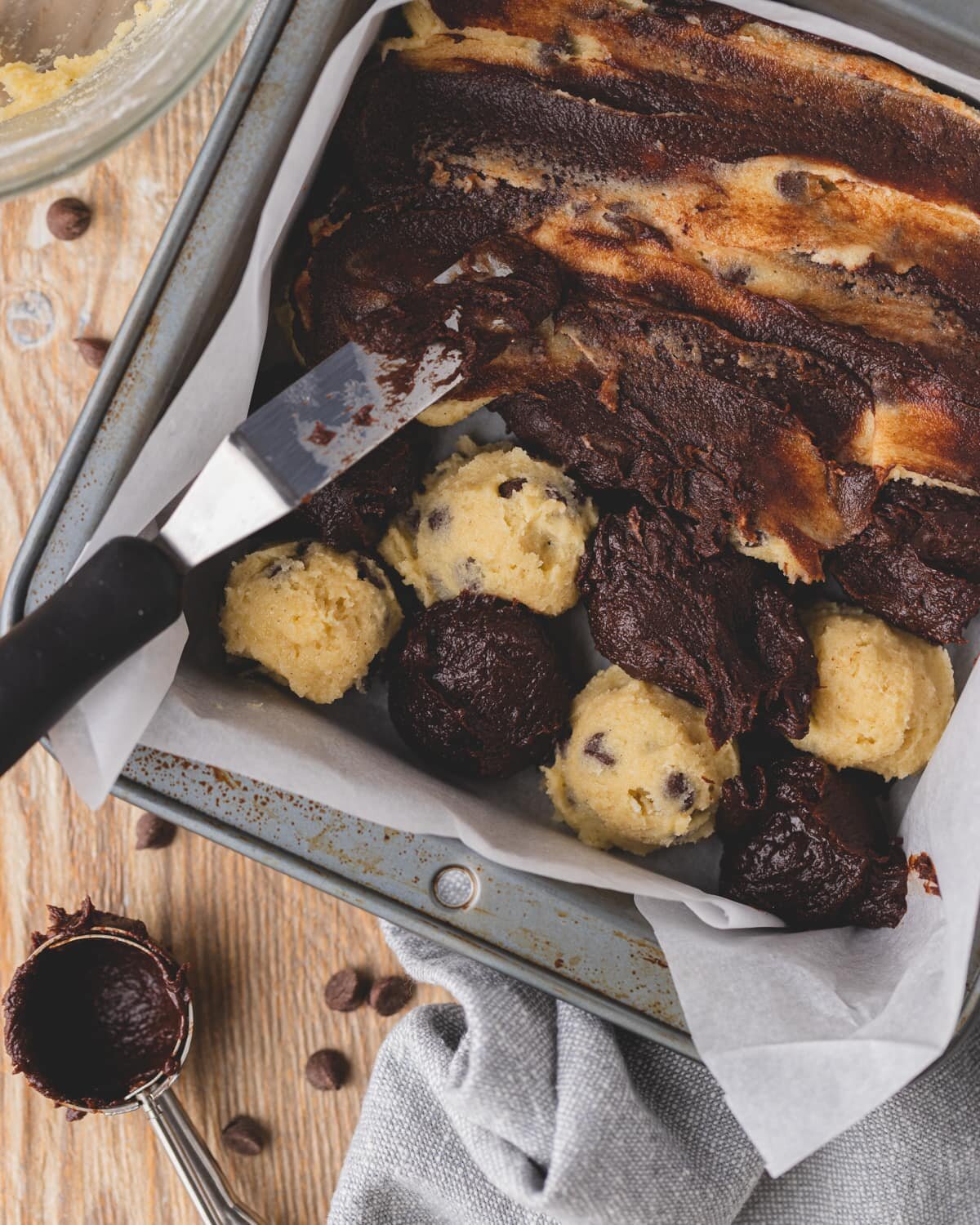 Alternating scoops of gluten free chocolate chip cookie dough and brownie batter in an 8x8 baking pan. A spreader is used to spread both doughs together before baking.
