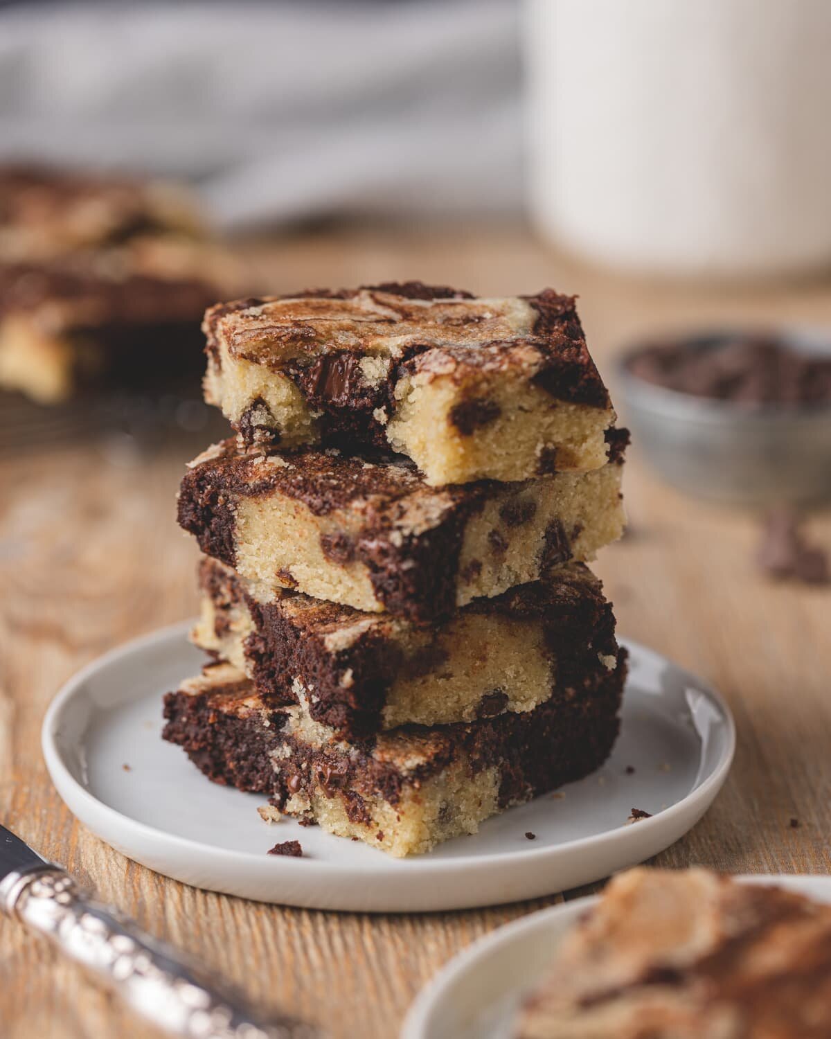 Four squares of gluten free cookie brownies stacked on top of a white plate - the top square has a bite missing and shows the melted chocolate chips inside.