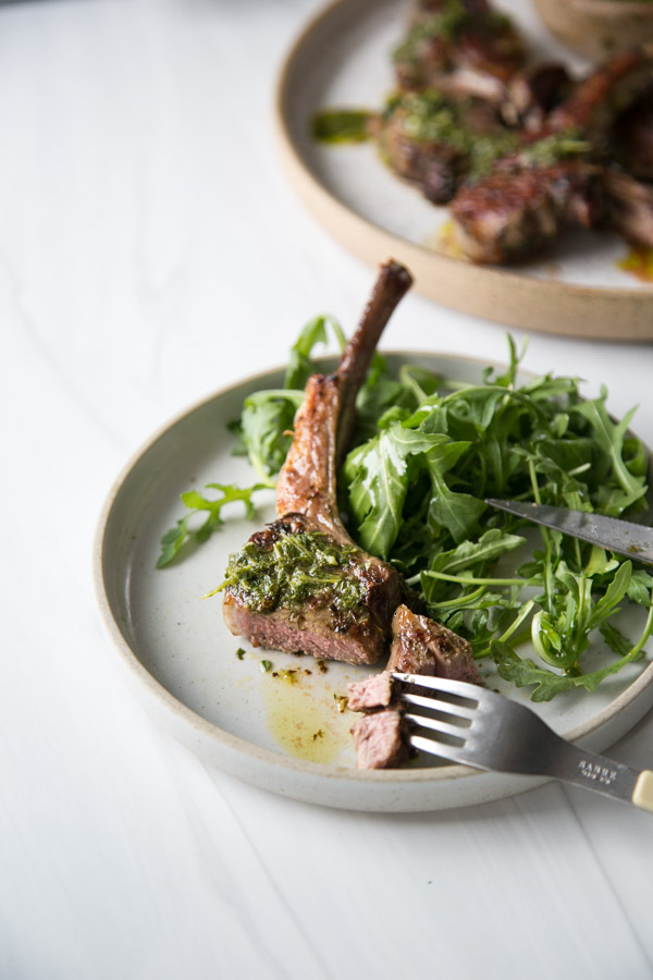 knife and fork cutting marinated lamb chops drizzled with mint sauce on a plate with arugula salad