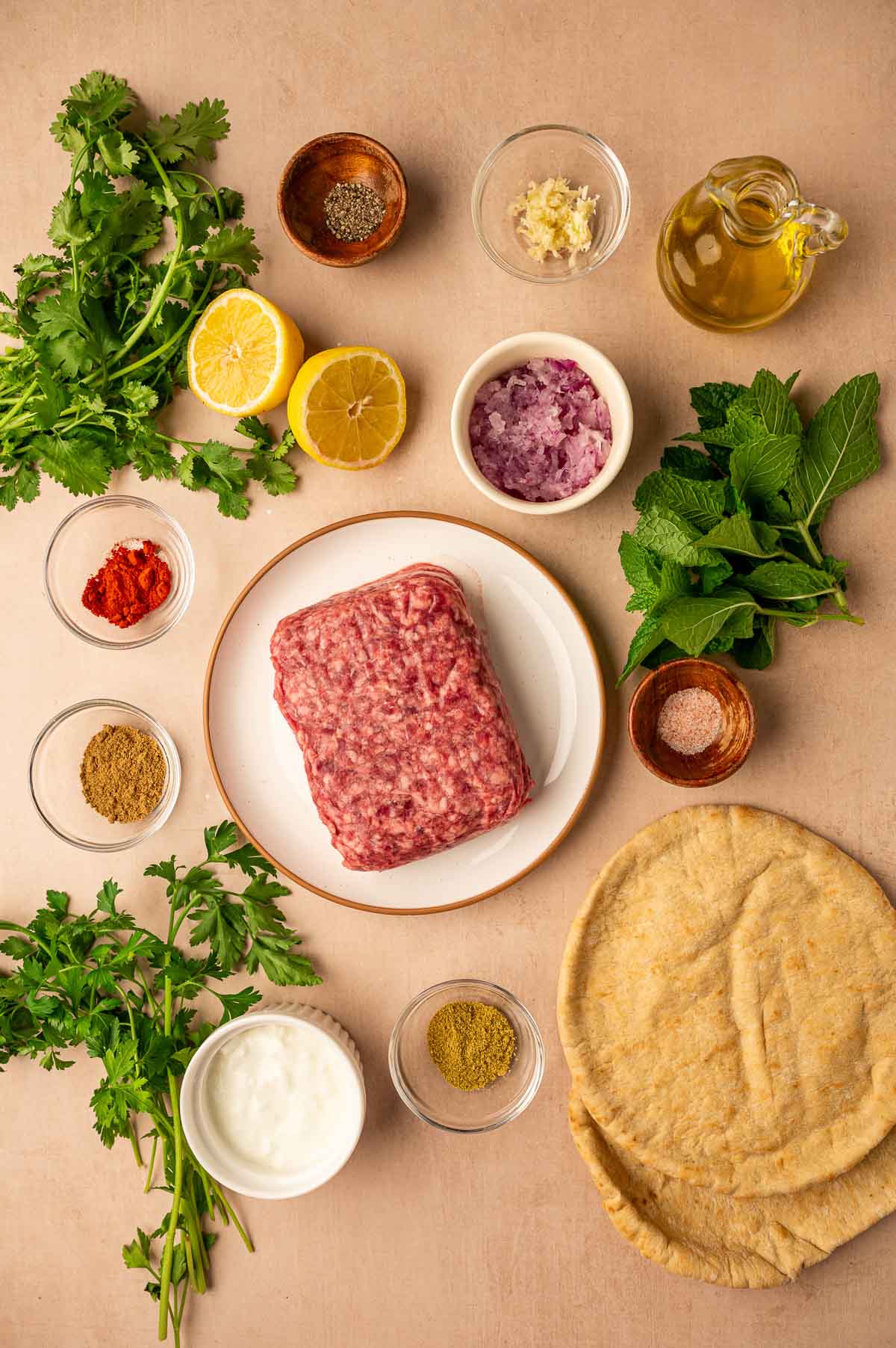 Raw ground lamb on a plate surrounded by pita bread, fresh herbs, lemon halves, spices, chopped onion, yogurt, garlic, olive oil, and seasoning bowls on a beige surface.