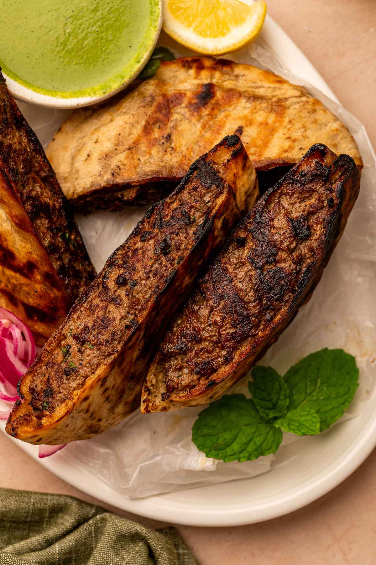 Two grilled lamb arayes on a plate with mint leaves, lemon, pickled onions, and a small bowl of green sauce.