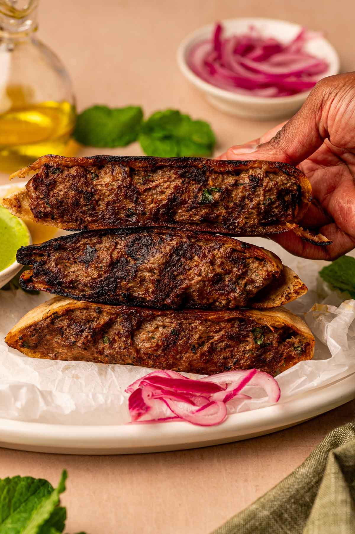 A hand holds a lamb arais, with pickled onions, mint leaves, and a bottle of oil in the background.