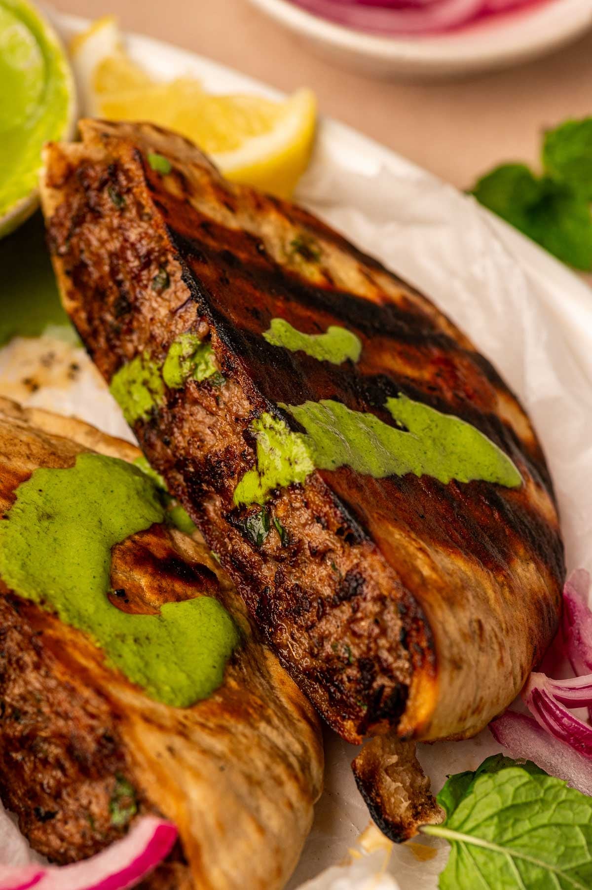Close-up of a grilled lamb arayes, topped with green sauce, served with lemon wedges and red onions.