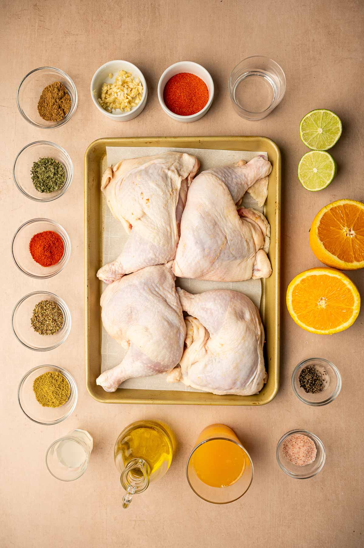 A tray with four raw chicken leg quarters is surrounded by bowls of spices, chopped garlic, liquids, halved citrus fruits, and bottles of oil and juice on a beige surface.
