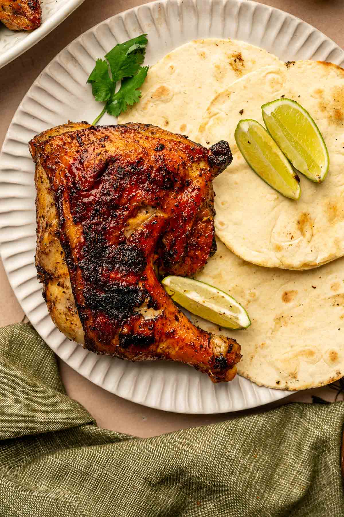 Grilled chicken leg quarter served with two corn tortillas, lime wedges, and a sprig of cilantro on a white plate.