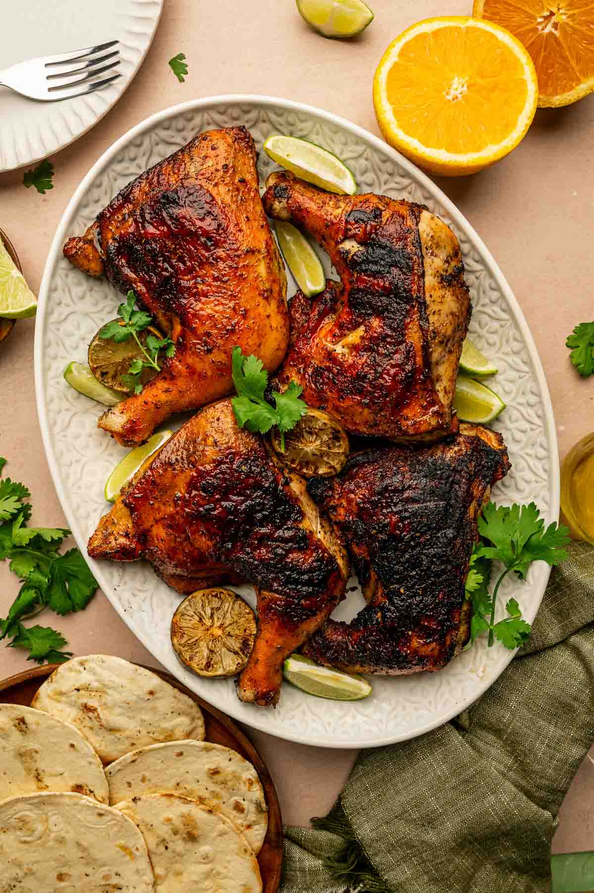 Grilled pollo asado garnished with lime wedges and cilantro on an oval platter, with tortillas, orange, and lime halves nearby.