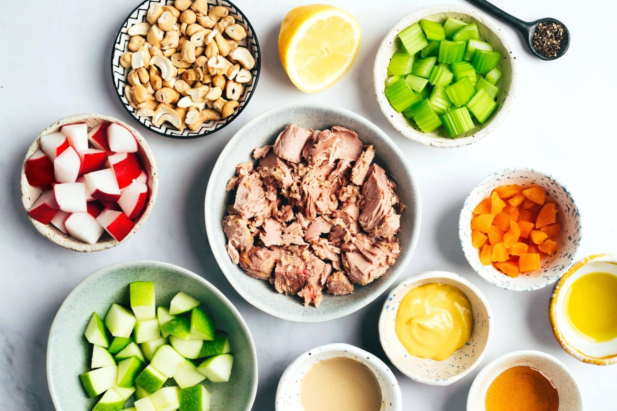 Ingredients for healthy tuna salad arranged on a white surface with canned tuna, celery, apples, radishes, cashews, lemon, and creamy dressing.