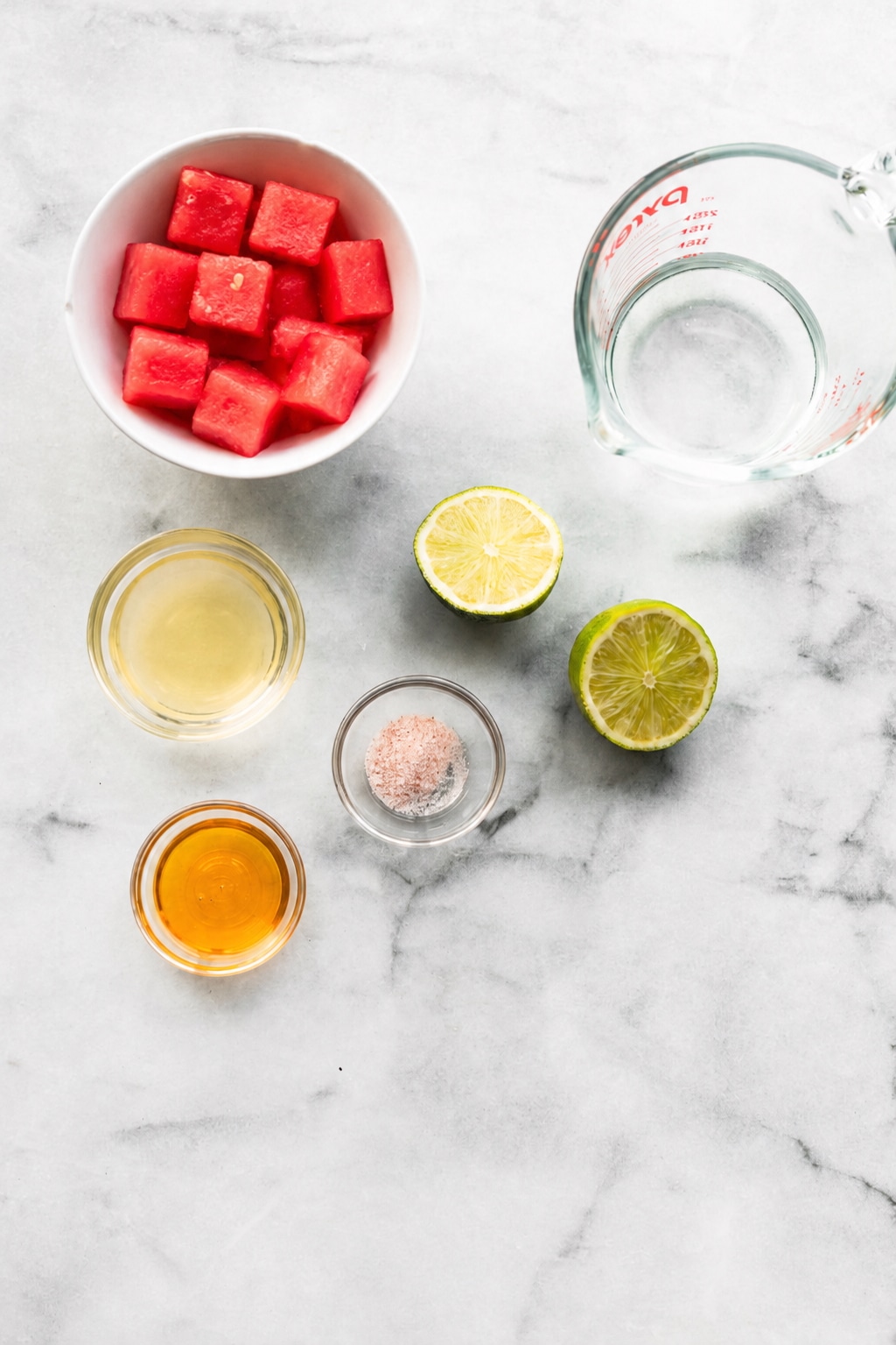 Ingredients for a homemade watermelon drink arranged on a marble surface, including watermelon, lime, sea salt, maple syrup, and water.