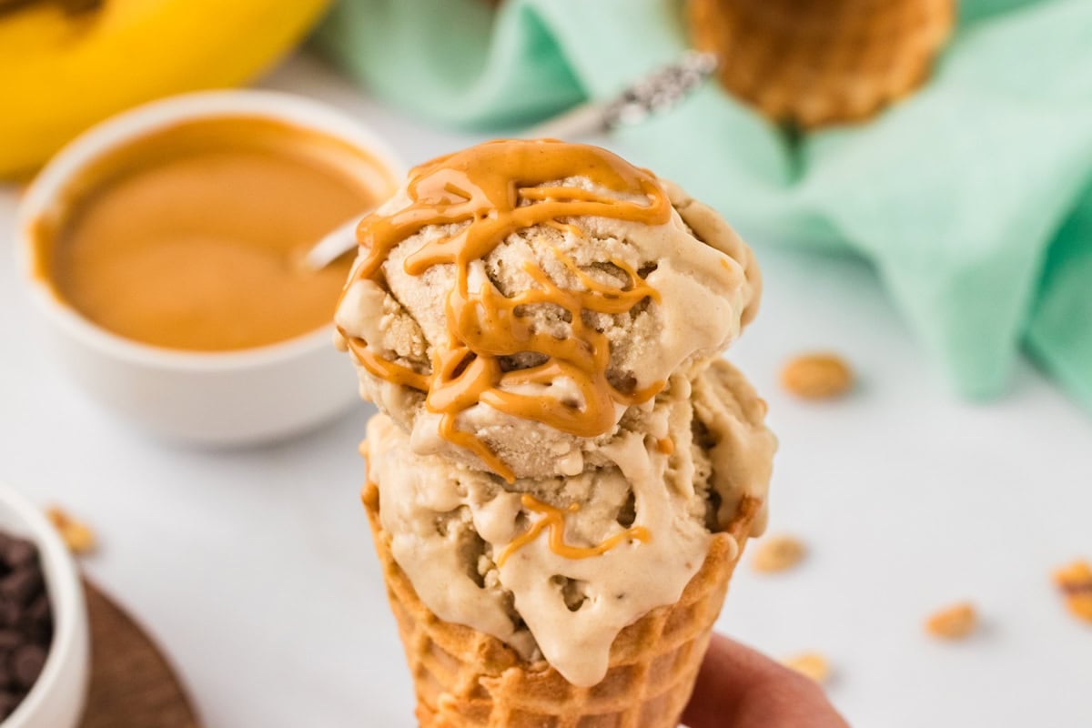 Hand holding a waffle cone with two scoops of banana peanut butter nice cream drizzled with melted peanut butter