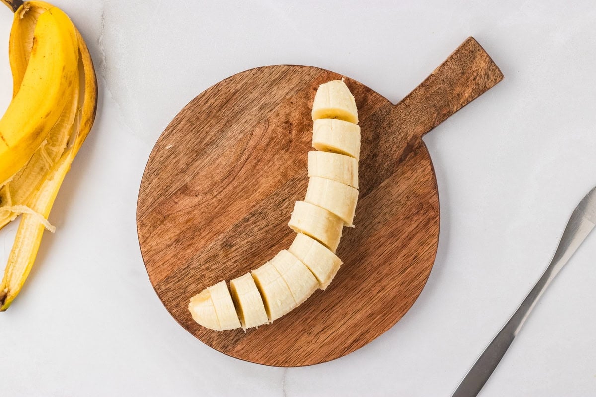 A ripe banana sliced into coins on a round wooden cutting board with the peel set to the side