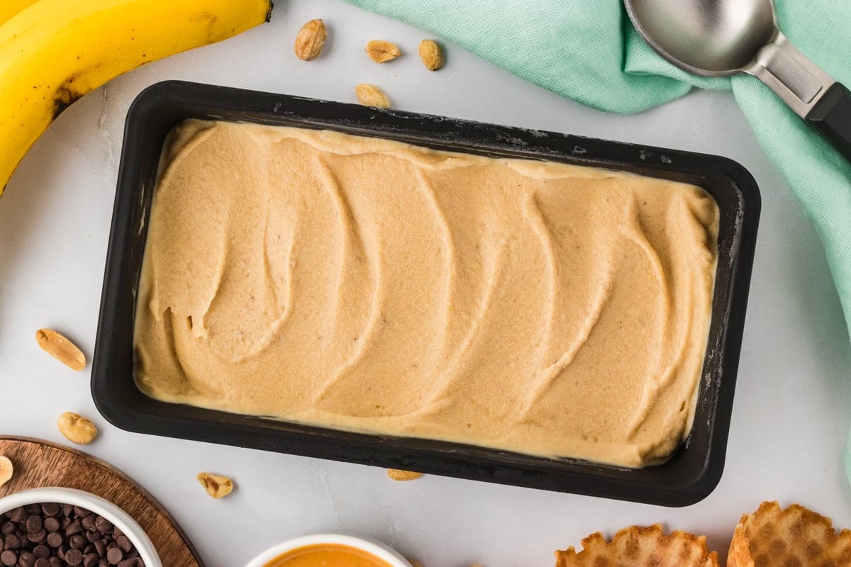 Overhead view of banana peanut butter nice cream in a black loaf pan surrounded by peanuts, chocolate chips, a waffle cone, and a banana