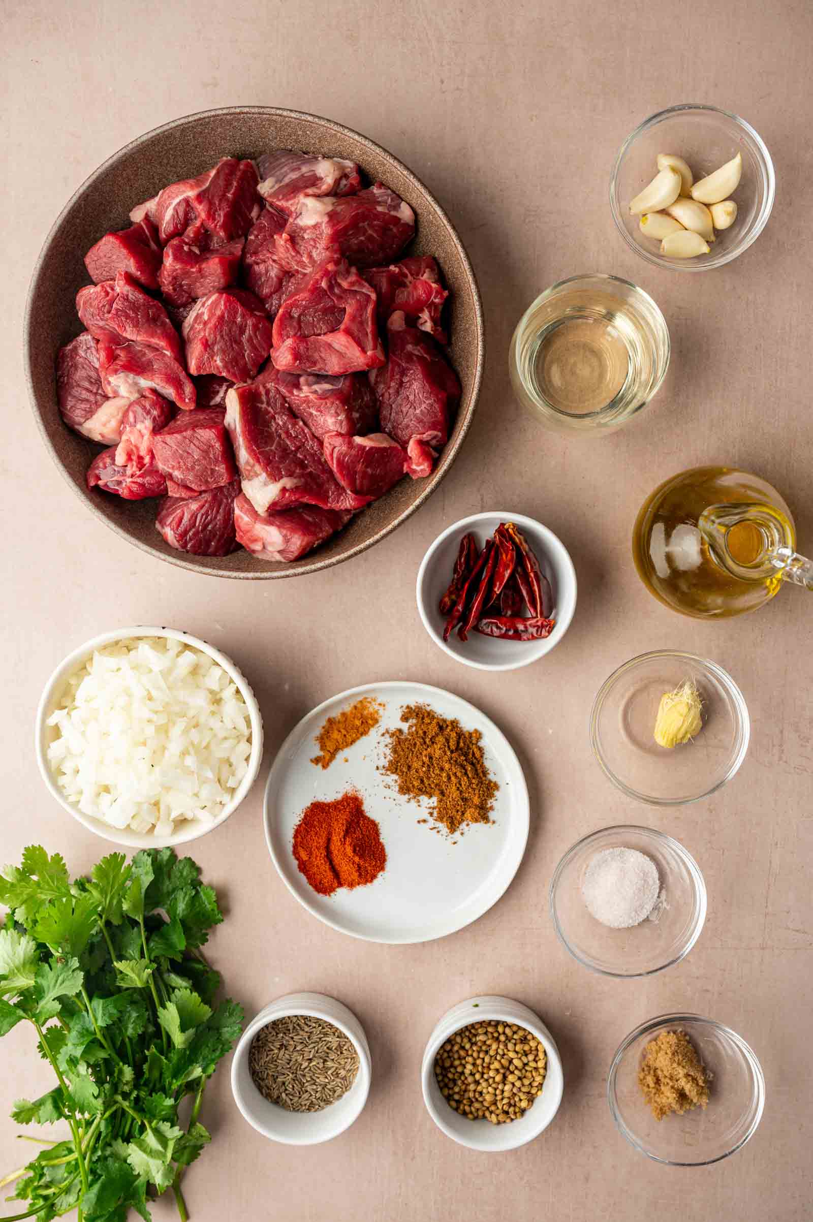 Overhead view of raw lamb chunks, chopped onion, various spices, garlic, fresh cilantro, oil, vinegar, and dried chilies arranged on a light surface.