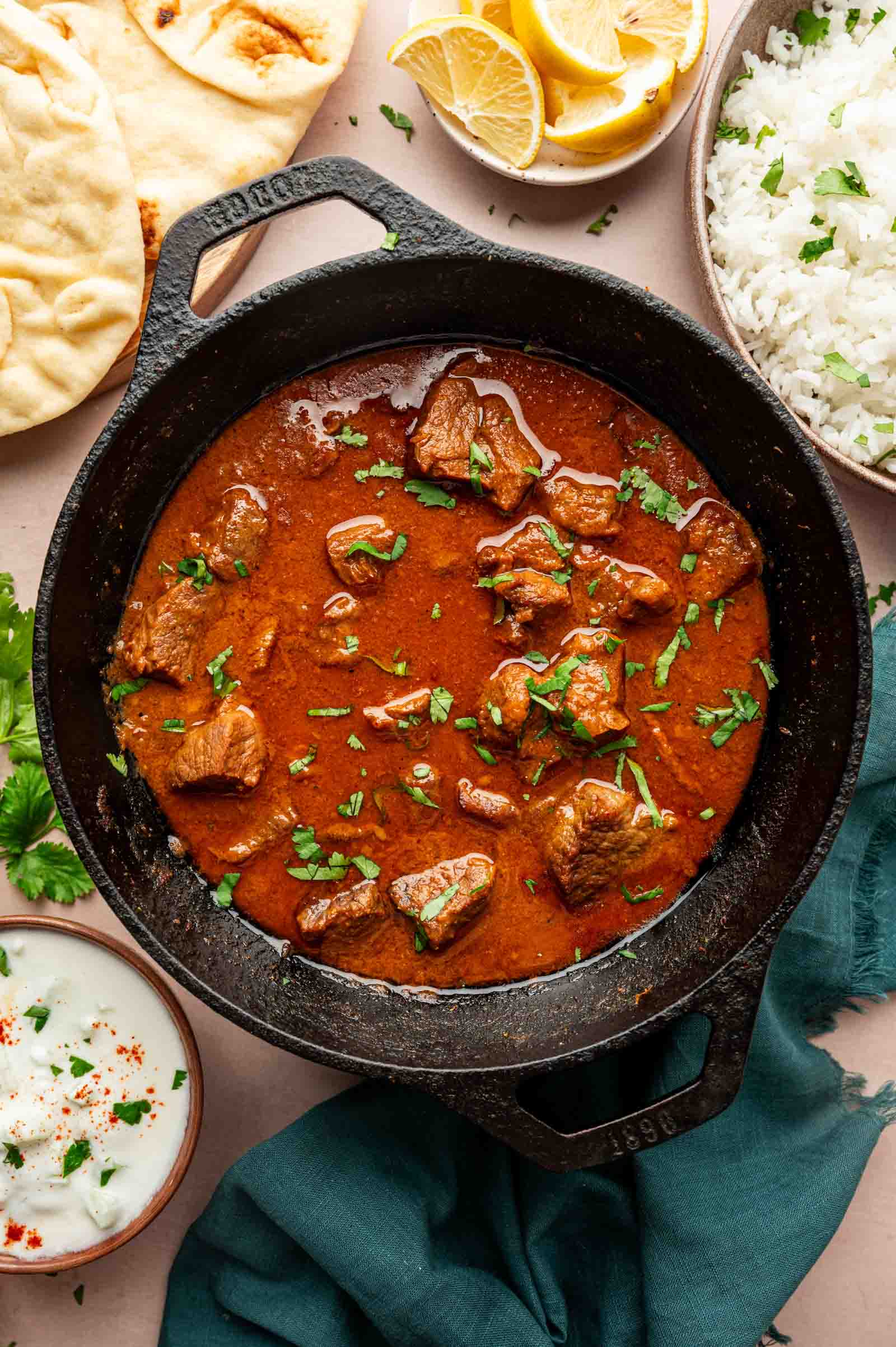 A cast iron pan filled with lamb vindaloo garnished with cilantro, surrounded by naan bread, lemon wedges, rice, and a bowl of raita sauce.