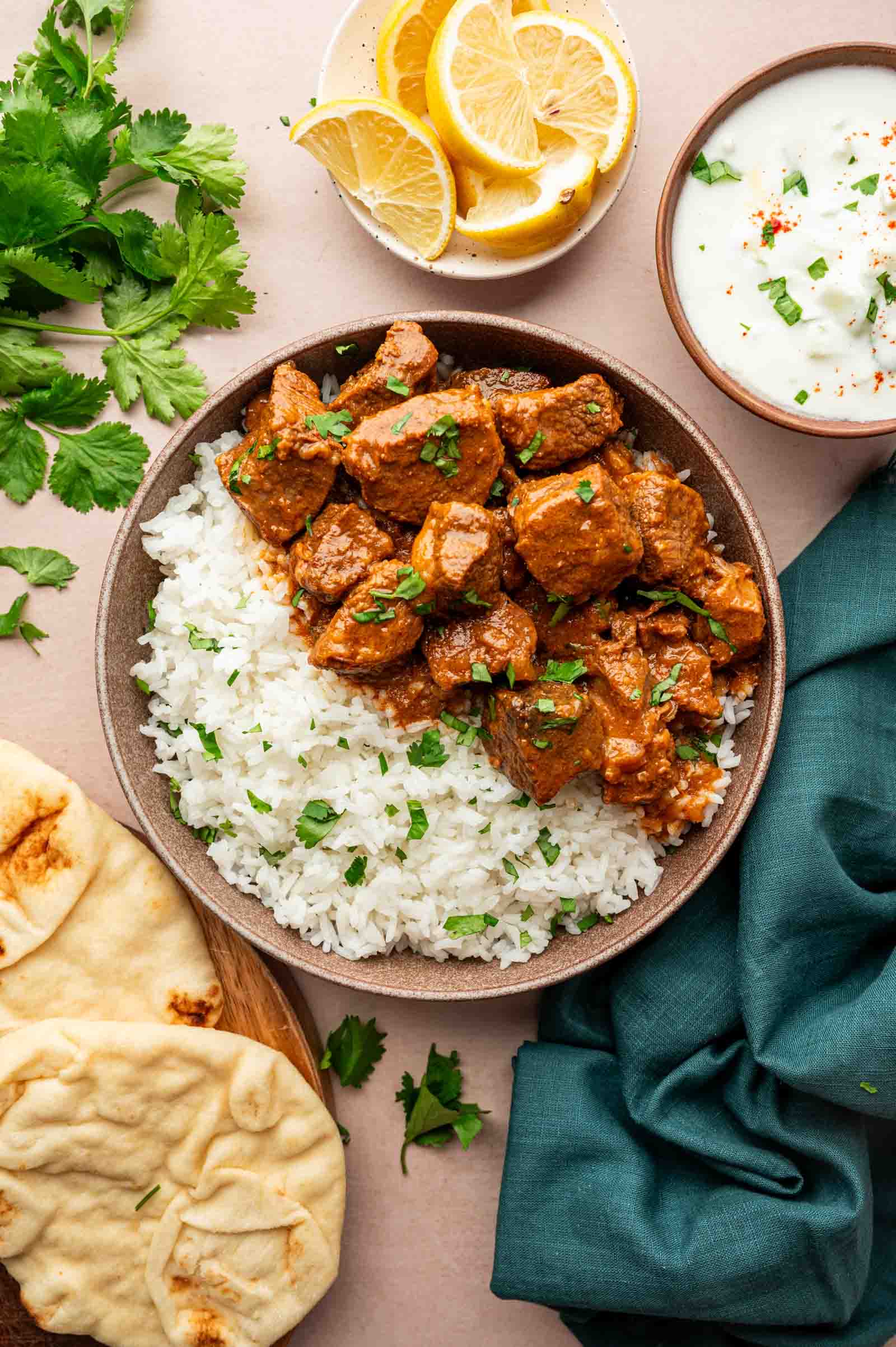 A bowl of white rice topped with chunks of lamb vindaloo, served with naan bread, lemon wedges, fresh cilantro, and a bowl of raita sauce on the side.