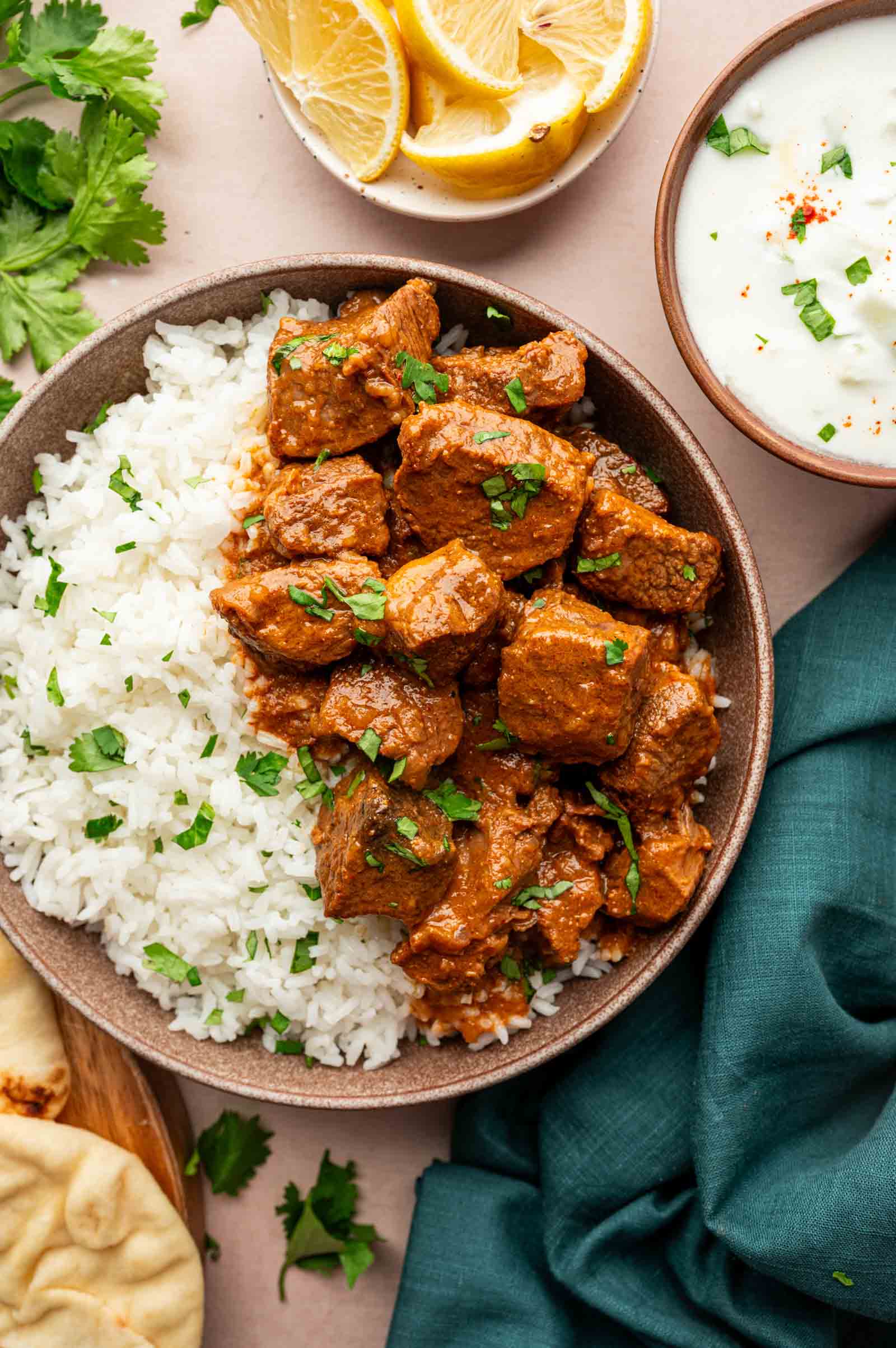 A bowl of white rice and lamb vindaloo garnished with cilantro, served with naan, lemon wedges, and a bowl of raita sauce on the side.