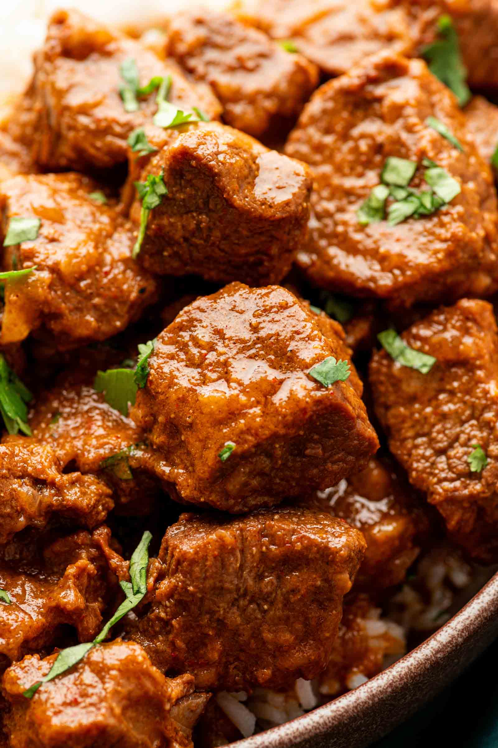 Close-up of lamb curry with chunks of meat in a rich, reddish-brown vindaloo sauce, garnished with chopped fresh cilantro, served over white rice.