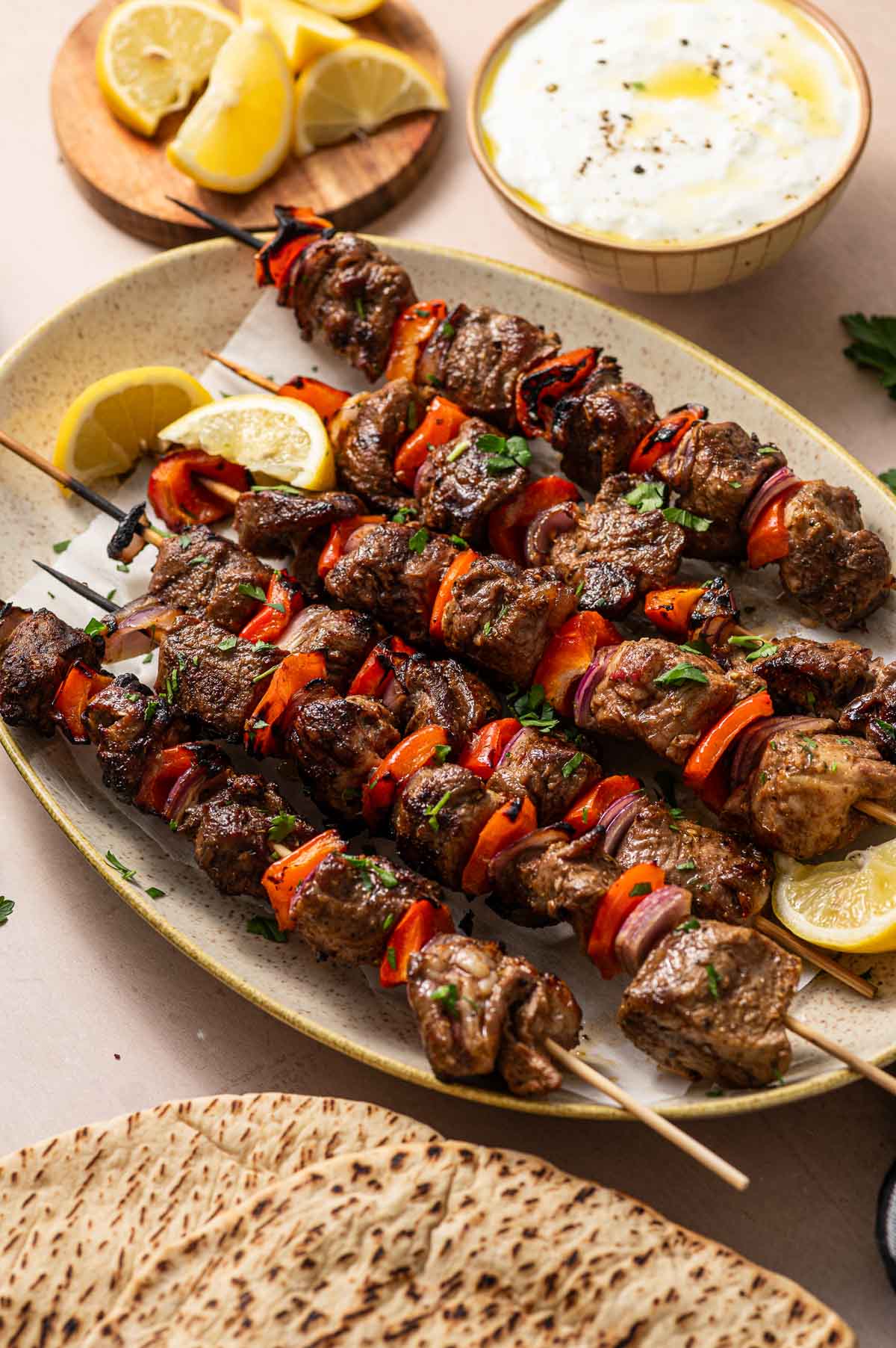 Grilled lamb and vegetable kabobs on a plate with lemon wedges, served alongside flatbread, lemon slices, and a bowl of tzatziki.