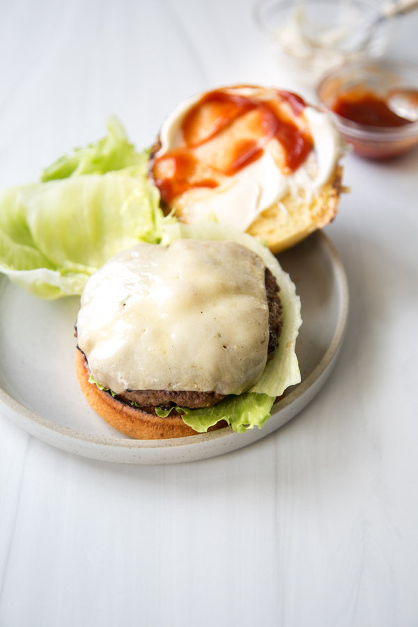 Open cheeseburger with lettuce and melted cheese on the patty, served on a gluten-free bun, with the top bun spread with mayonnaise and ketchup, placed on a round plate on a white surface.
