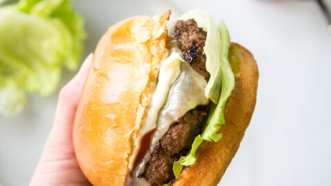 A hand holds up a gluten-free cheeseburger with lettuce and a toasted bun; a plate with lettuce and a jar of ketchup with a spoon are blurred in the background.