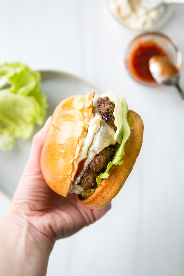 A hand holds up a gluten-free cheeseburger with lettuce and a toasted bun; a plate with lettuce and a jar of ketchup with a spoon are blurred in the background.