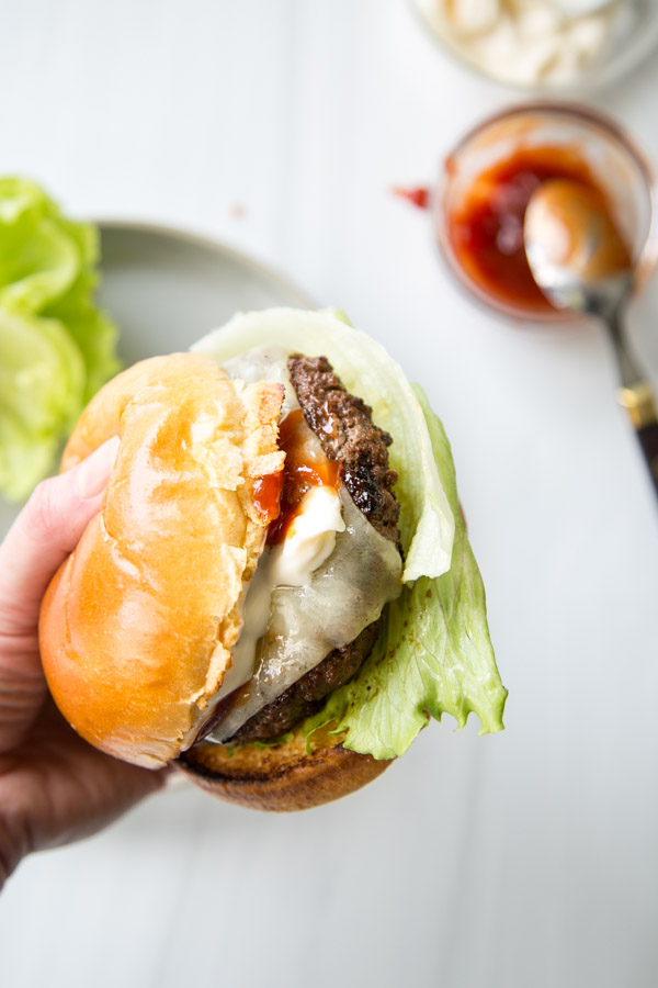 A hand holding a cheeseburger with lettuce, melted cheese, mayonnaise, and a shiny bun. In the background, there is a plate with lettuce and a jar of ketchup with a spoon next to a gluten-free blueberry cobbler.