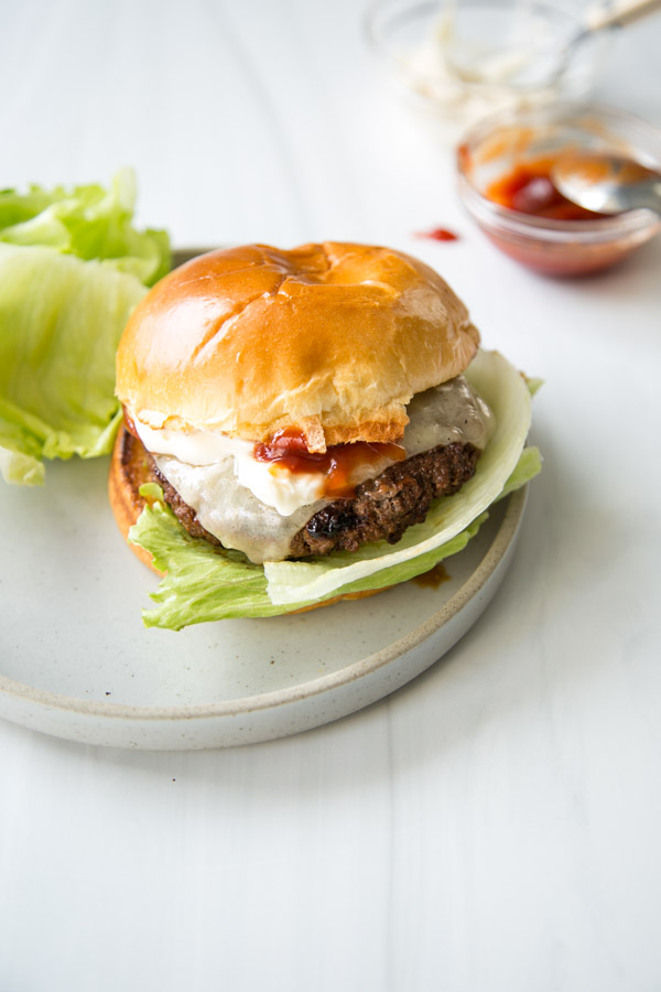 A gluten-free cheeseburger with lettuce, a slice of cheese, and ketchup in a brioche bun sits on a plate, with extra lettuce and a bowl of ketchup in the background.