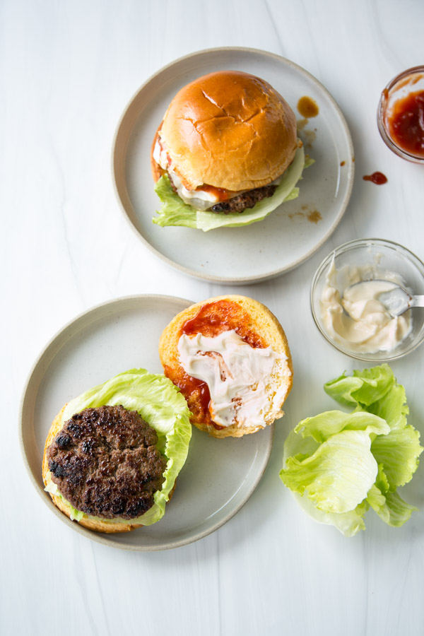 Two plates with hamburgers: one assembled and one open-faced. The open burger sits on a gluten-free bun with a beef patty, lettuce, mayonnaise, and ketchup. Nearby are a bowl of mayonnaise, ketchup jar, and fresh lettuce on a white surface.